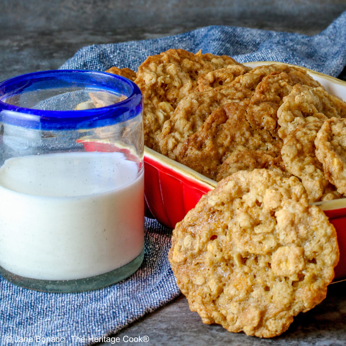 Golden brown Crunchy Oatmeal Cookies are standing upright in a small red baking dish, leaning against each other, with a glass of milk beside it and a single cookie propped up against the front of the dish. All sitting on a blue cloth with a gray background behind © 2026 Jane Bonacci, The Heritage Cook.