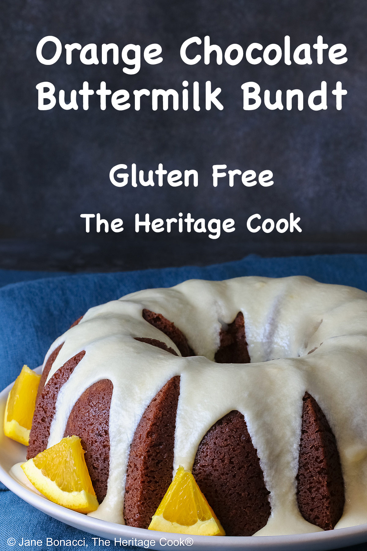 Whole chocolate/orange bundt cake with cascading orange flavored glaze and fresh orange slices around the exterior on a white plate, sometimes on its own, sometimes with a slice cut out and on a plate in front, on a blue cloth with a dark gray background © 2026 Jane Bonacci, The Heritage Cook.