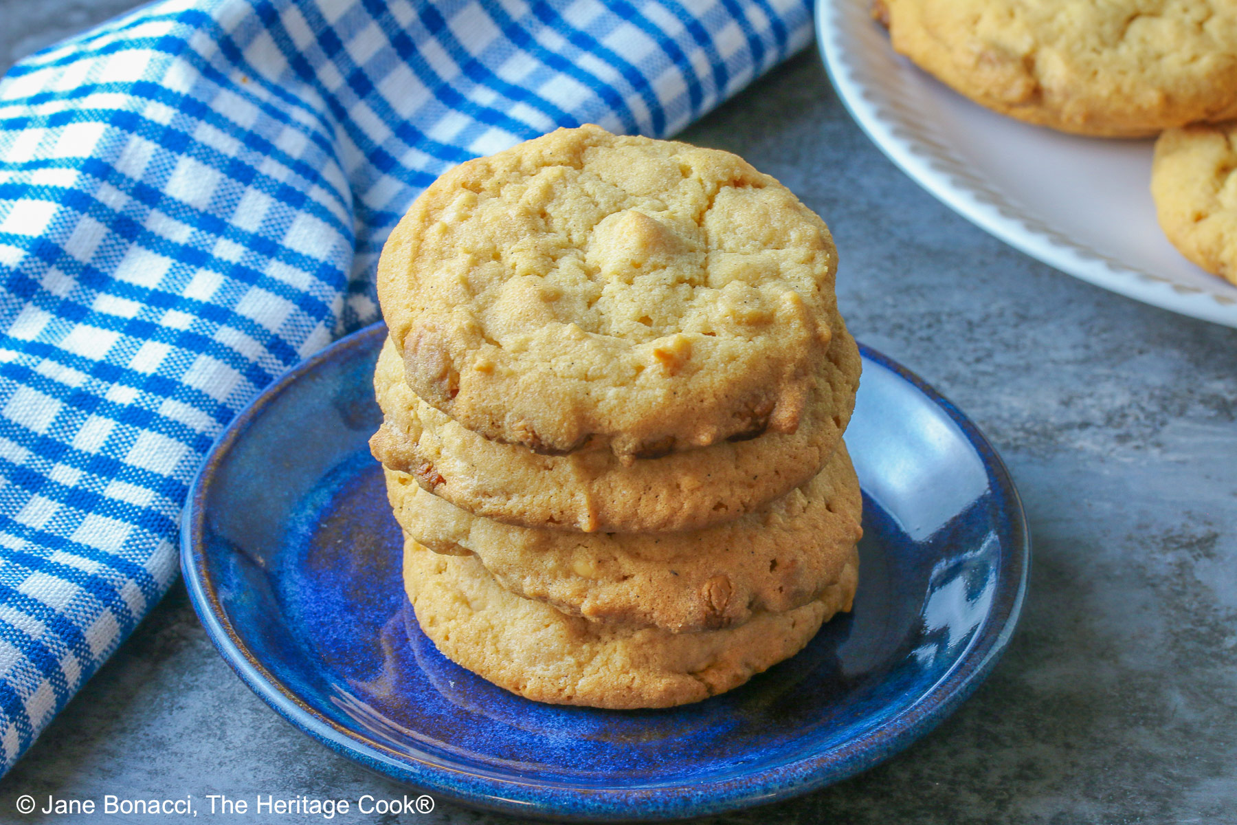 A stack of Peanut Butter White Chocolate Chip Cookies on a small blue plate with a white plate piled with more cookies behind, sitting on a gray background, some with a blue checked cloth around them © 2026 Jane Bonacci, The Heritage Cook.