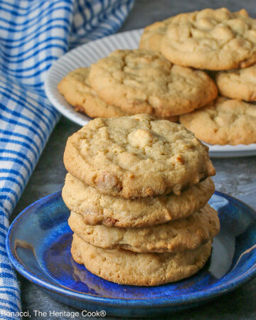 A stack of Peanut Butter White Chocolate Chip Cookies on a small blue plate with a white plate piled with more cookies behind, sitting on a gray background, some with a blue checked cloth around them © 2026 Jane Bonacci, The Heritage Cook.