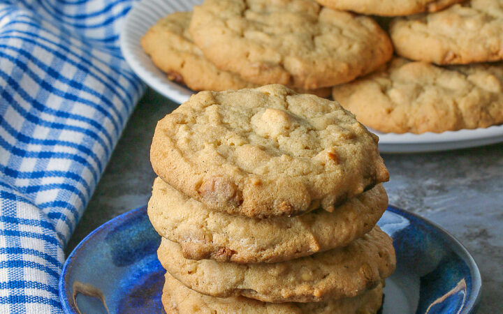 A stack of Peanut Butter White Chocolate Chip Cookies on a small blue plate with a white plate piled with more cookies behind, sitting on a gray background, some with a blue checked cloth around them © 2026 Jane Bonacci, The Heritage Cook.