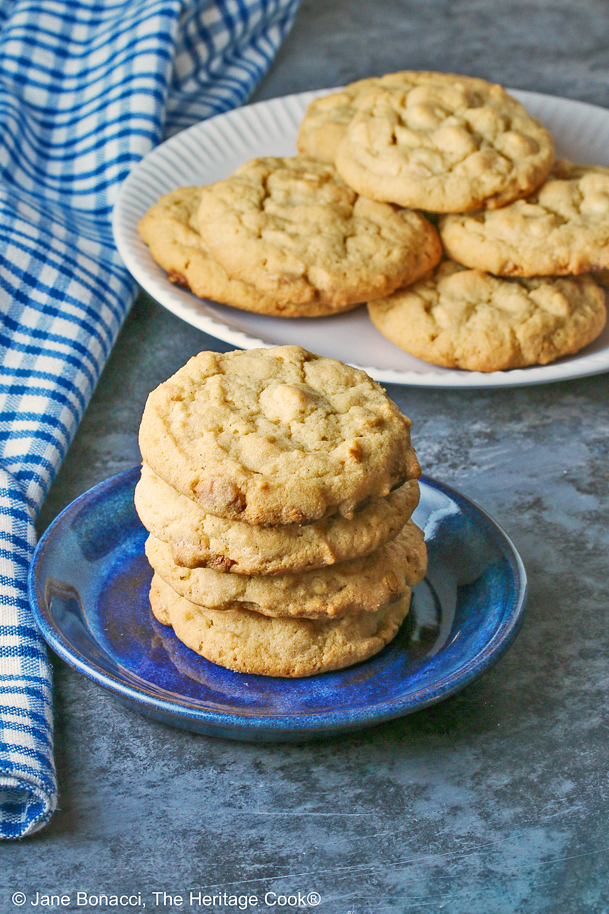 A stack of Peanut Butter White Chocolate Chip Cookies on a small blue plate with a white plate piled with more cookies behind, sitting on a gray background, some with a blue checked cloth around them © 2026 Jane Bonacci, The Heritage Cook.