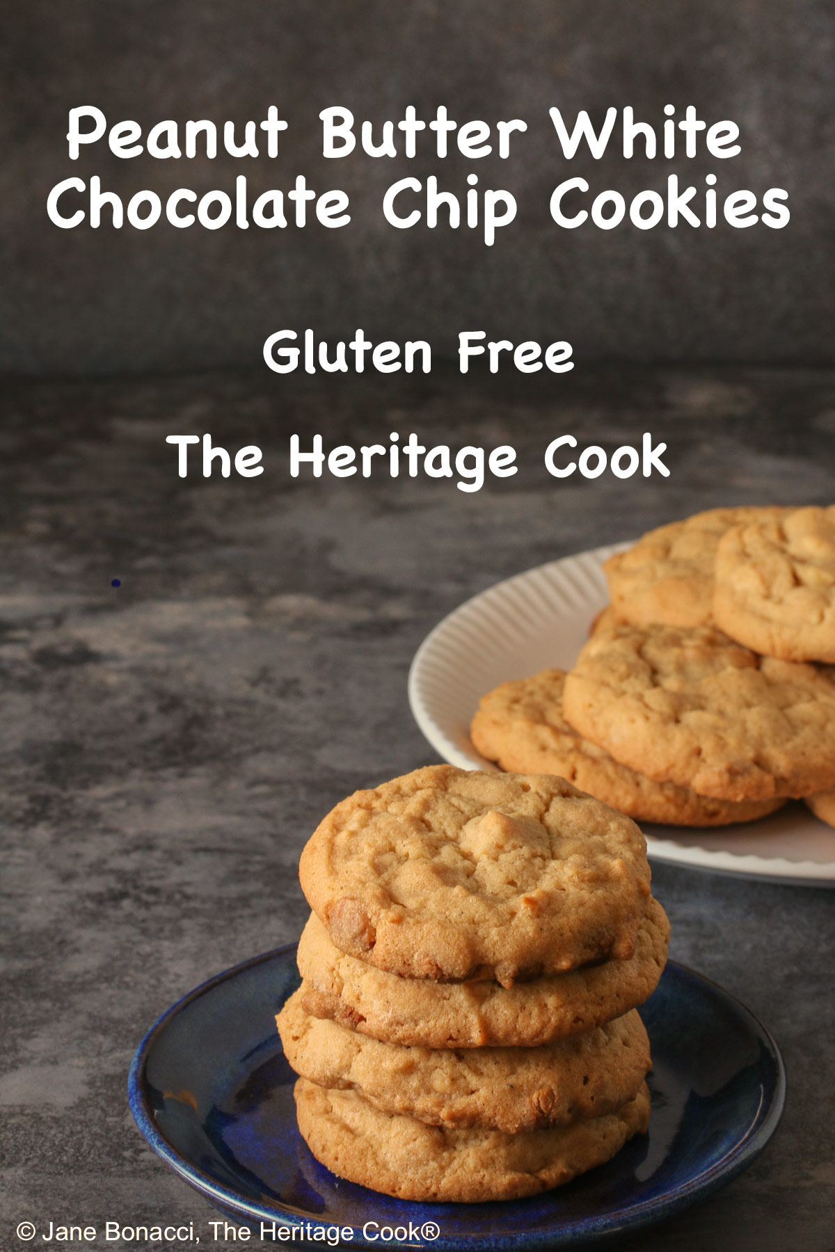 A stack of Peanut Butter White Chocolate Chip Cookies on a small blue plate with a white plate piled with more cookies behind, sitting on a gray background, some with a blue checked cloth around them © 2026 Jane Bonacci, The Heritage Cook.