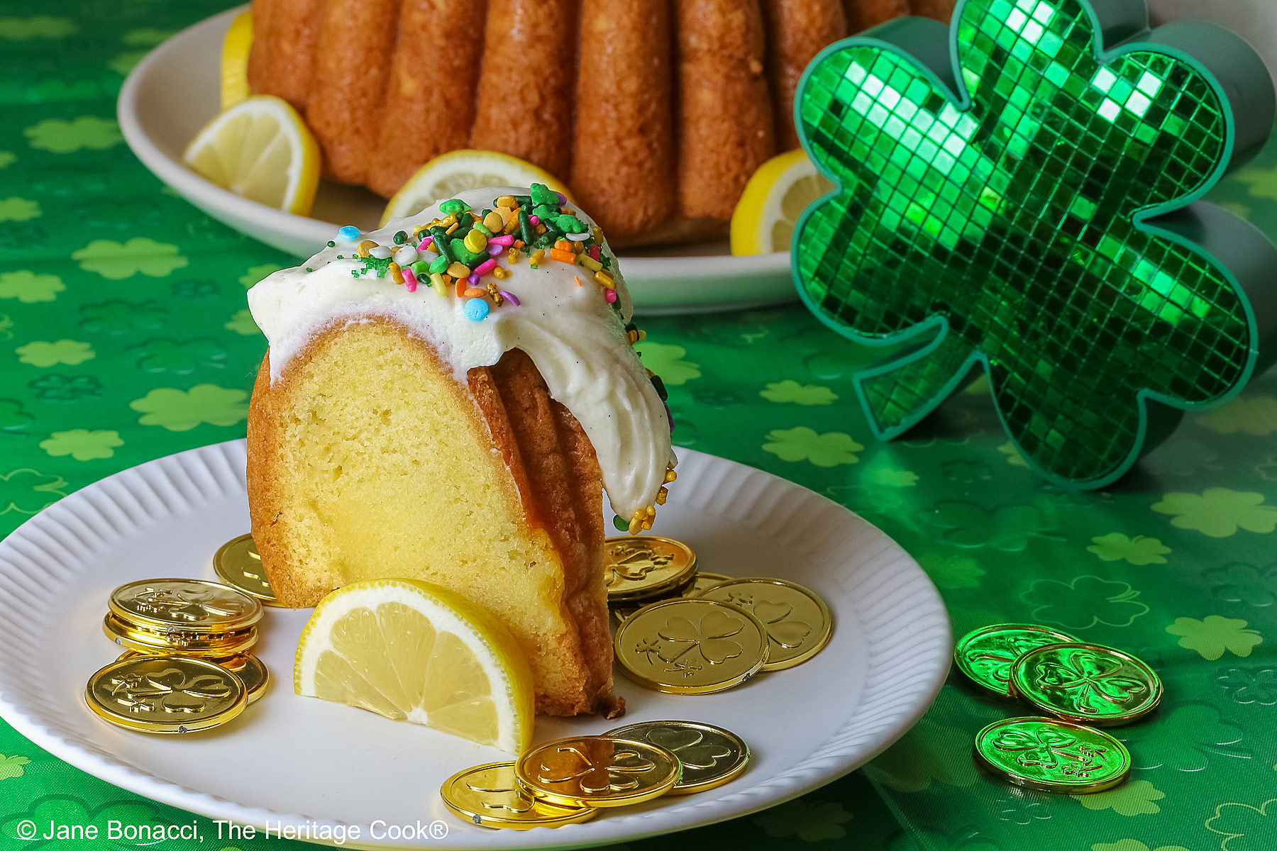 Whole Golden Lemon Bundt Cake on a white plate with lemon slices around the base, topped with fluffy white chocolate butter cream frosting and colorful sprinkles for St. Patrick’s Day; a slice of cake in front surrounded by gold coins from the pot ‘o gold of the leprechauns and a bright green cloth © 2026 Jane Bonacci, The Heritage Cook.