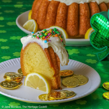 Whole Golden Lemon Bundt Cake on a white plate with lemon slices around the base, topped with fluffy white chocolate butter cream frosting and colorful sprinkles for St. Patrick’s Day; a slice of cake in front surrounded by gold coins from the pot ‘o gold of the leprechauns and a bright green cloth © 2026 Jane Bonacci, The Heritage Cook.