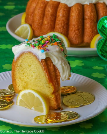 Whole Golden Lemon Bundt Cake on a white plate with lemon slices around the base, topped with fluffy white chocolate butter cream frosting and colorful sprinkles for St. Patrick’s Day; a slice of cake in front surrounded by gold coins from the pot ‘o gold of the leprechauns and a bright green cloth © 2026 Jane Bonacci, The Heritage Cook.