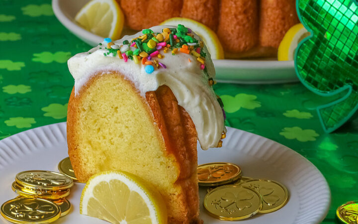 Whole Golden Lemon Bundt Cake on a white plate with lemon slices around the base, topped with fluffy white chocolate butter cream frosting and colorful sprinkles for St. Patrick’s Day; a slice of cake in front surrounded by gold coins from the pot ‘o gold of the leprechauns and a bright green cloth © 2026 Jane Bonacci, The Heritage Cook.