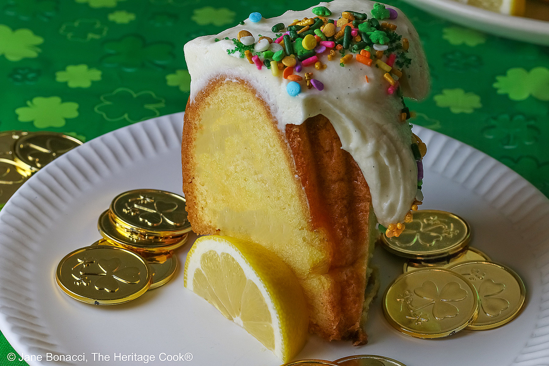 Whole Golden Lemon Bundt Cake on a white plate with lemon slices around the base, topped with fluffy white chocolate butter cream frosting and colorful sprinkles for St. Patrick’s Day; a slice of cake in front surrounded by gold coins from the pot ‘o gold of the leprechauns and a bright green cloth © 2026 Jane Bonacci, The Heritage Cook.