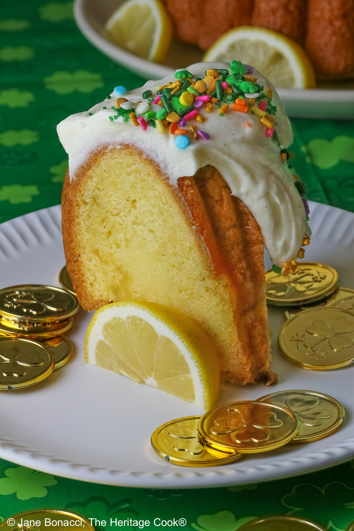 Whole Golden Lemon Bundt Cake on a white plate with lemon slices around the base, topped with fluffy white chocolate butter cream frosting and colorful sprinkles for St. Patrick’s Day; a slice of cake in front surrounded by gold coins from the pot ‘o gold of the leprechauns and a bright green cloth © 2026 Jane Bonacci, The Heritage Cook.