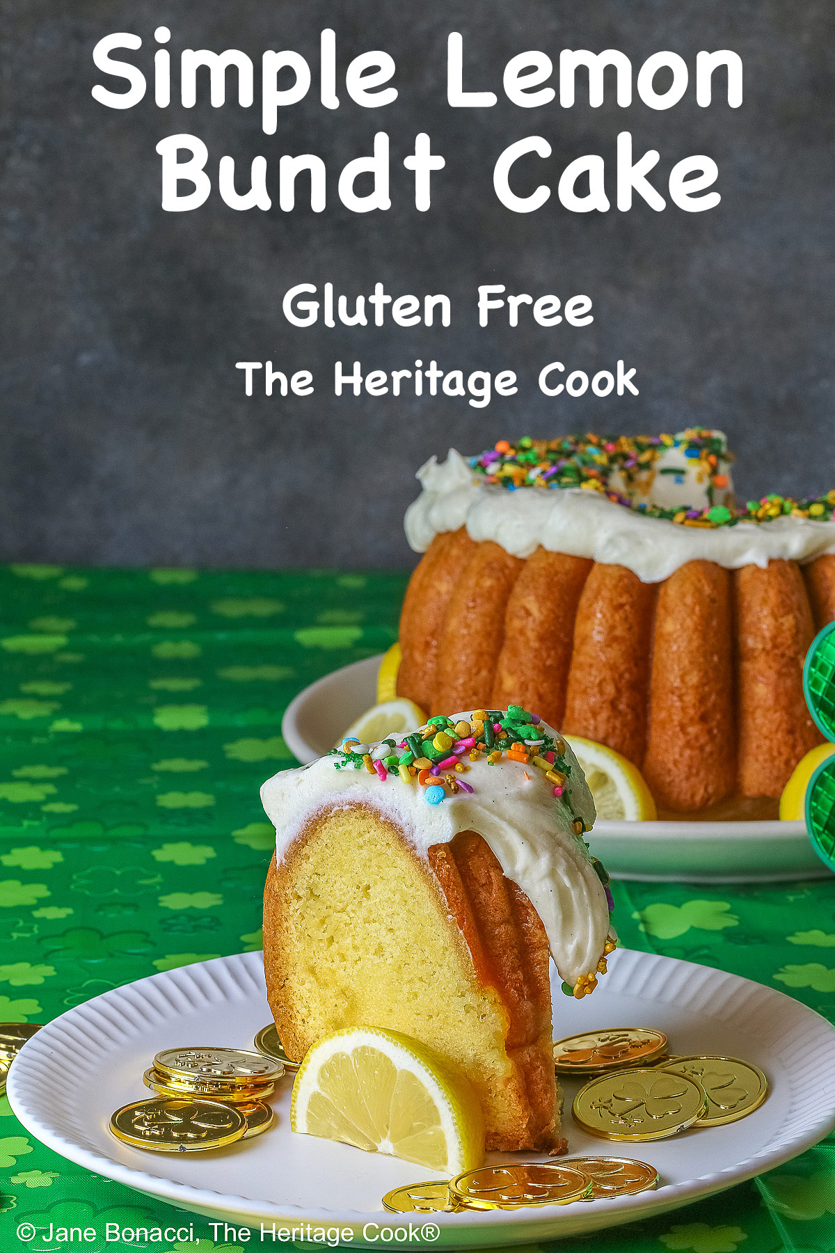 Whole Golden Lemon Bundt Cake on a white plate with lemon slices around the base, topped with fluffy white chocolate butter cream frosting and colorful sprinkles for St. Patrick’s Day; a slice of cake in front surrounded by gold coins from the pot ‘o gold of the leprechauns and a bright green cloth © 2026 Jane Bonacci, The Heritage Cook.