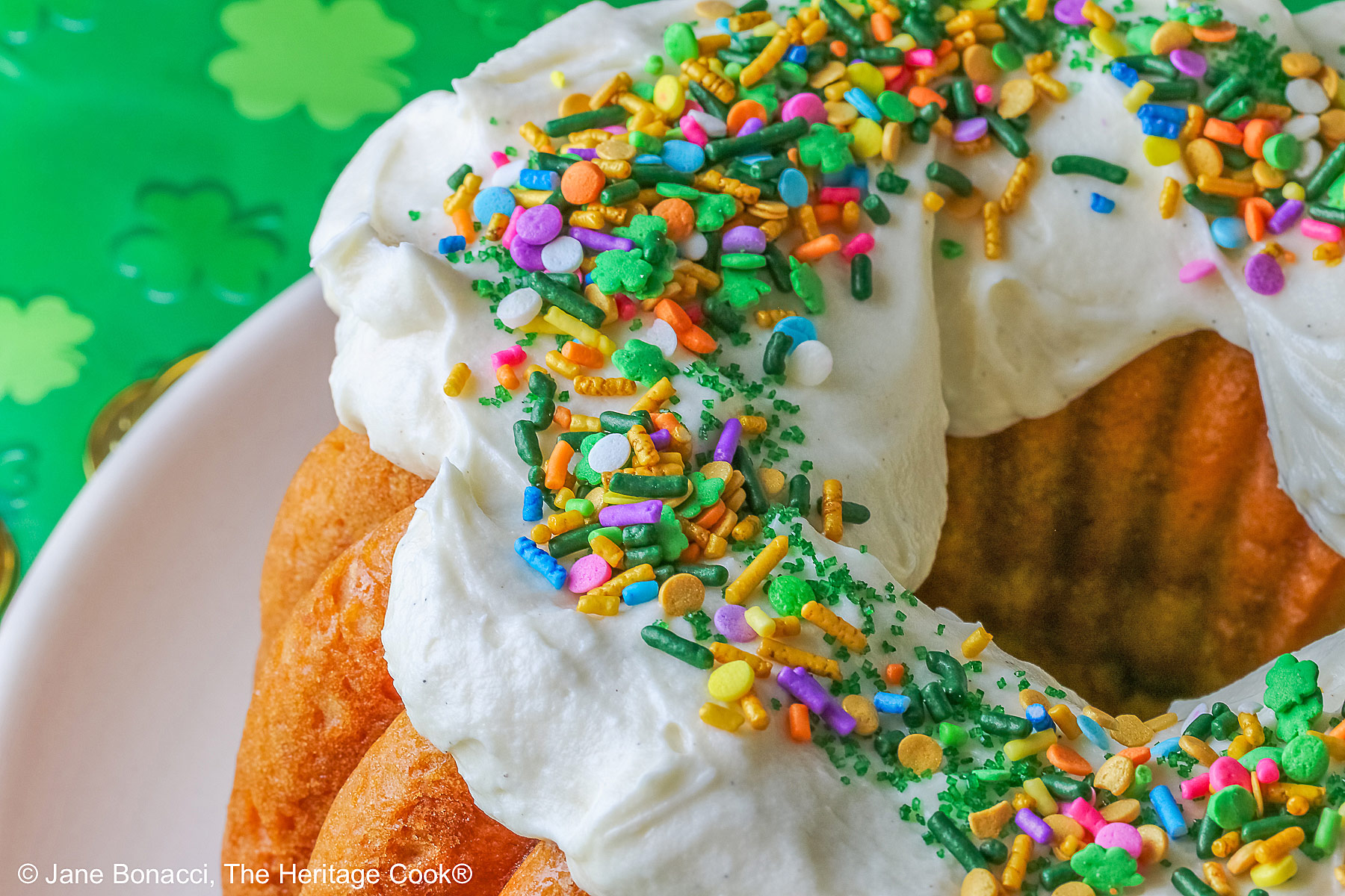 Whole Golden Lemon Bundt Cake on a white plate with lemon slices around the base, topped with fluffy white chocolate butter cream frosting and colorful sprinkles for St. Patrick’s Day; a slice of cake in front surrounded by gold coins from the pot ‘o gold of the leprechauns and a bright green cloth © 2026 Jane Bonacci, The Heritage Cook.