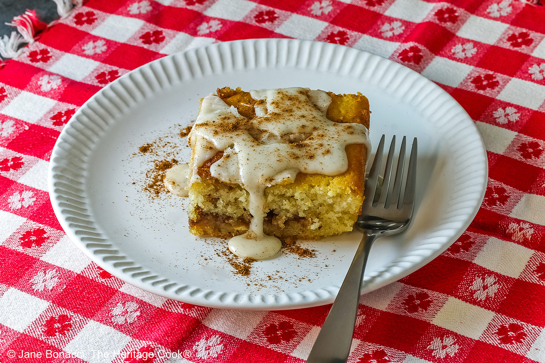 Large square slice of Cinnamon Roll Cake on a white plate with ridges around the outer edge and a fork on the side; there is a glaze over the top that is dripping down onto the plate which is sitting on a red and white checked cloth © 2026 Jane Bonacci, The Heritage Cook.