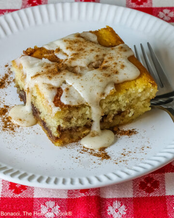 Large square slice of Cinnamon Roll Cake on a white plate with ridges around the outer edge and a fork on the side; there is a glaze over the top that is dripping down onto the plate which is sitting on a red and white checked cloth © 2026 Jane Bonacci, The Heritage Cook.