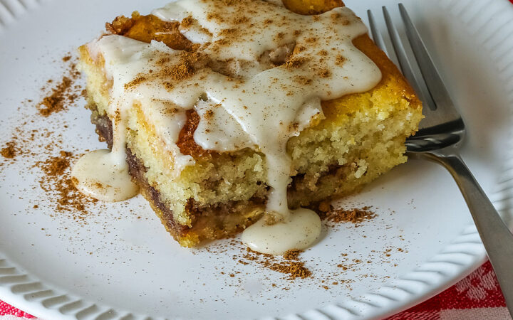 Large square slice of Cinnamon Roll Cake on a white plate with ridges around the outer edge and a fork on the side; there is a glaze over the top that is dripping down onto the plate which is sitting on a red and white checked cloth © 2026 Jane Bonacci, The Heritage Cook.