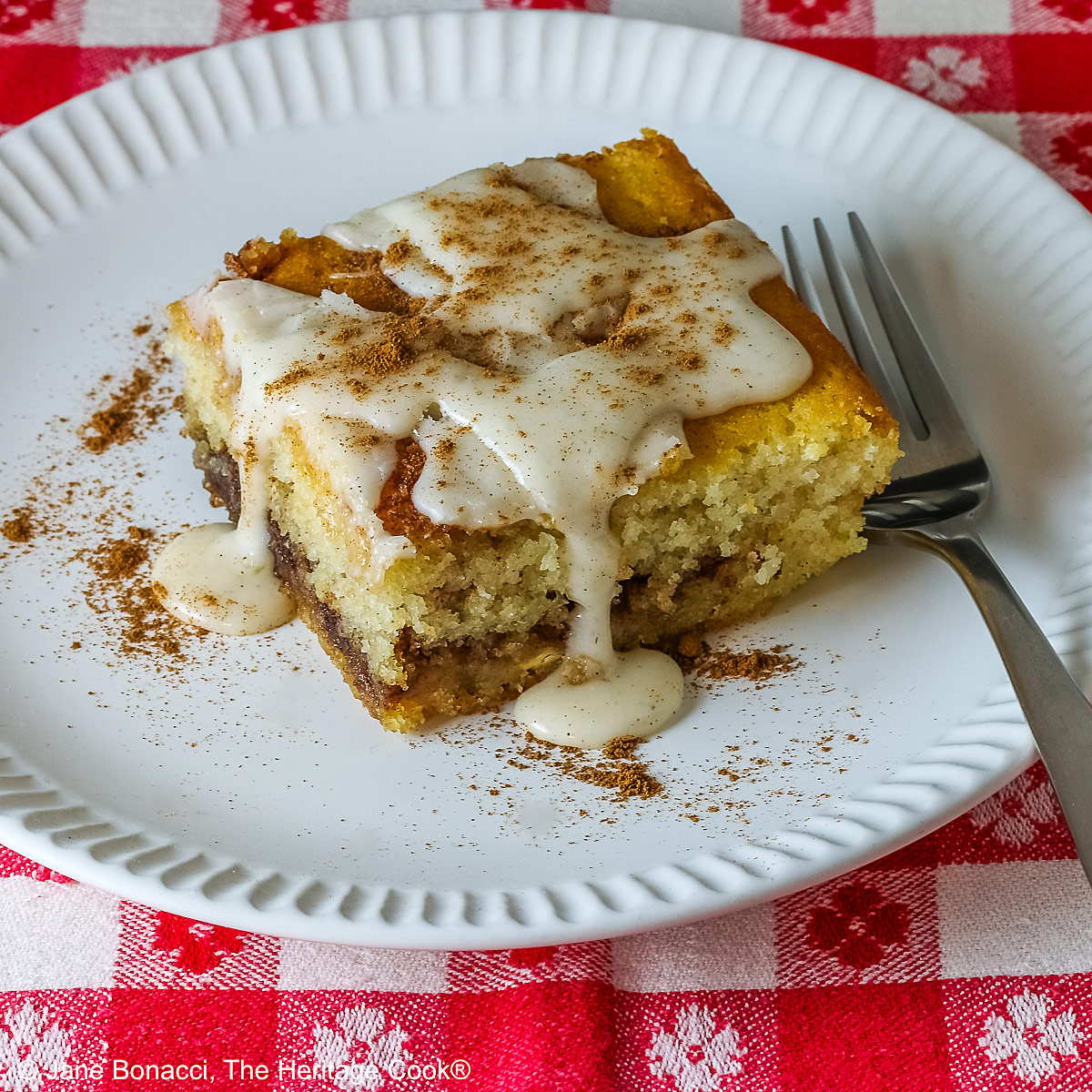 Large square slice of Cinnamon Roll Cake on a white plate with ridges around the outer edge and a fork on the side; there is a glaze over the top that is dripping down onto the plate which is sitting on a red and white checked cloth © 2026 Jane Bonacci, The Heritage Cook.
