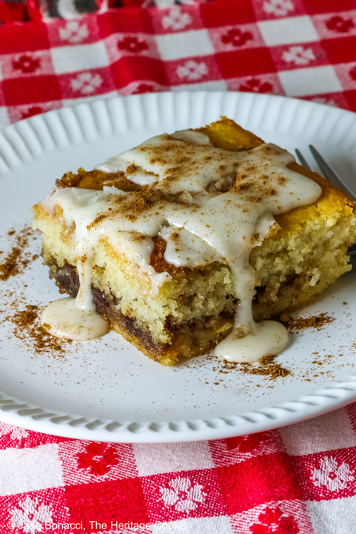 Large square slice of Cinnamon Roll Cake on a white plate with ridges around the outer edge and a fork on the side; there is a glaze over the top that is dripping down onto the plate which is sitting on a red and white checked cloth © 2026 Jane Bonacci, The Heritage Cook.