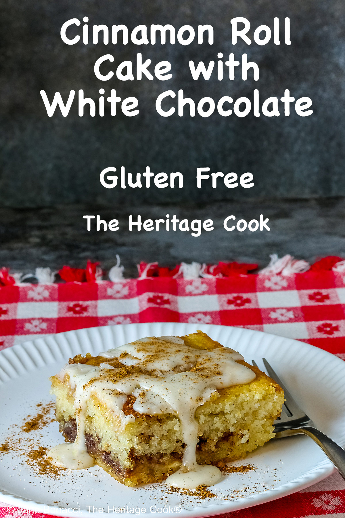 Large square slice of Cinnamon Roll Cake on a white plate with ridges around the outer edge and a fork on the side; there is a glaze over the top that is dripping down onto the plate which is sitting on a red and white checked cloth © 2026 Jane Bonacci, The Heritage Cook.