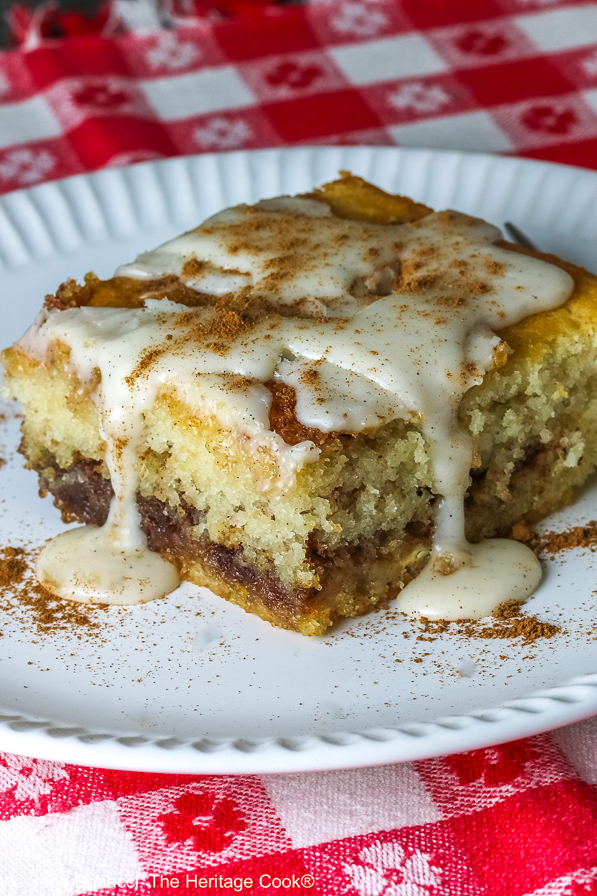 Large square slice of Cinnamon Roll Cake on a white plate with ridges around the outer edge and a fork on the side; there is a glaze over the top that is dripping down onto the plate which is sitting on a red and white checked cloth © 2026 Jane Bonacci, The Heritage Cook.