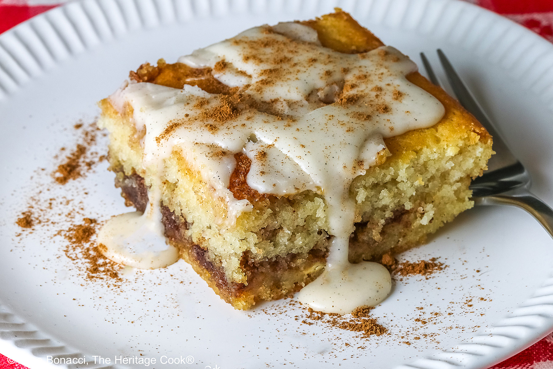 Large square slice of Cinnamon Roll Cake on a white plate with ridges around the outer edge and a fork on the side; there is a glaze over the top that is dripping down onto the plate which is sitting on a red and white checked cloth © 2026 Jane Bonacci, The Heritage Cook.