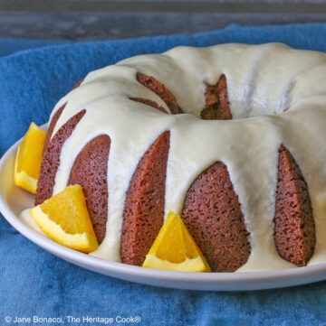 Whole chocolate/orange bundt cake with cascading orange flavored glaze and fresh orange slices around the exterior on a white plate, sometimes on its own, sometimes with a slice cut out and on a plate in front, on a blue cloth with a dark gray background © 2026 Jane Bonacci, The Heritage Cook.