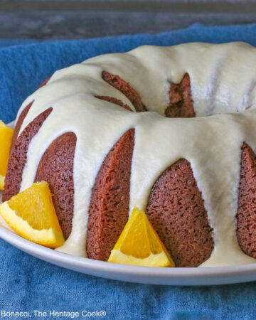 Whole chocolate/orange bundt cake with cascading orange flavored glaze and fresh orange slices around the exterior on a white plate, sometimes on its own, sometimes with a slice cut out and on a plate in front, on a blue cloth with a dark gray background © 2026 Jane Bonacci, The Heritage Cook.