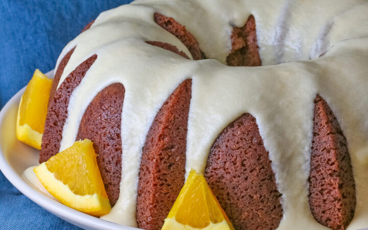 Whole chocolate/orange bundt cake with cascading orange flavored glaze and fresh orange slices around the exterior on a white plate, sometimes on its own, sometimes with a slice cut out and on a plate in front, on a blue cloth with a dark gray background © 2026 Jane Bonacci, The Heritage Cook.