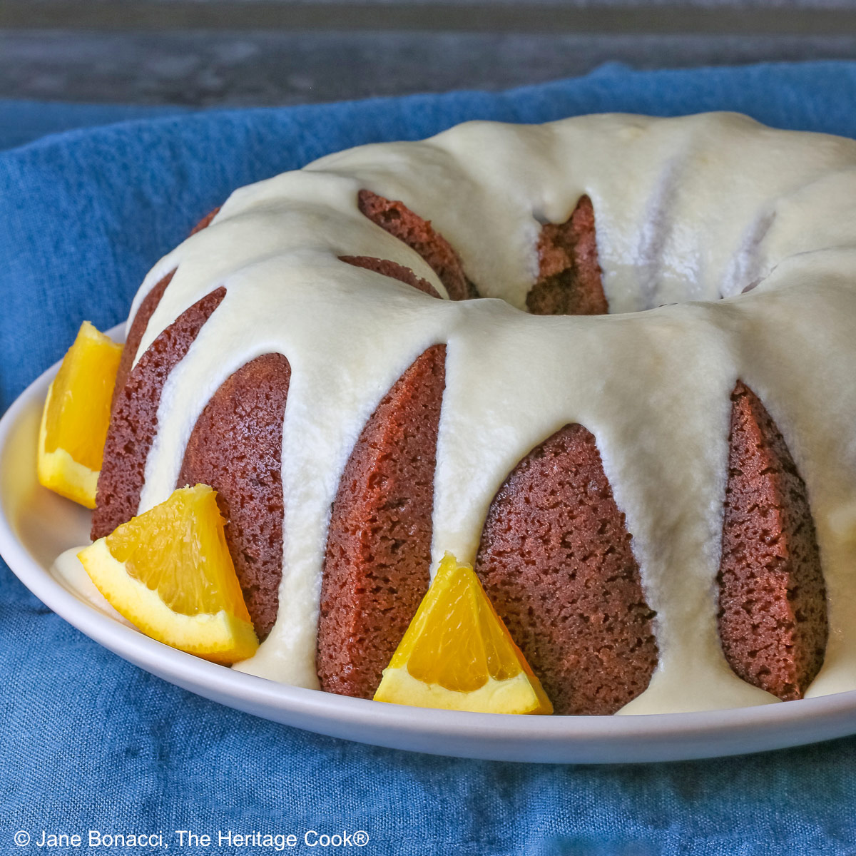 Whole chocolate/orange bundt cake with cascading orange flavored glaze and fresh orange slices around the exterior on a white plate, sometimes on its own, sometimes with a slice cut out and on a plate in front, on a blue cloth with a dark gray background © 2026 Jane Bonacci, The Heritage Cook.