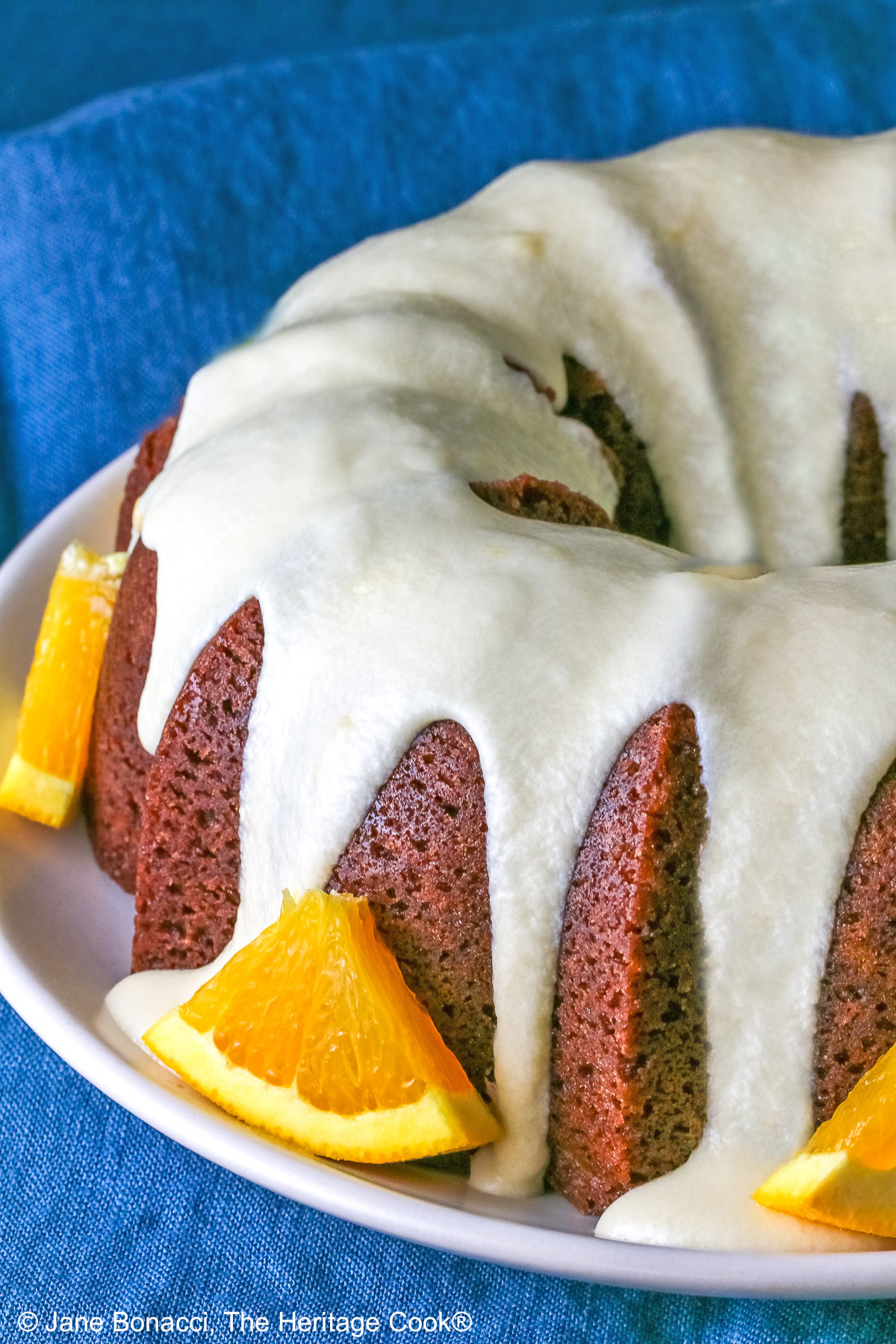 Whole chocolate/orange bundt cake with cascading orange flavored glaze and fresh orange slices around the exterior on a white plate, sometimes on its own, sometimes with a slice cut out and on a plate in front, on a blue cloth with a dark gray background © 2026 Jane Bonacci, The Heritage Cook.