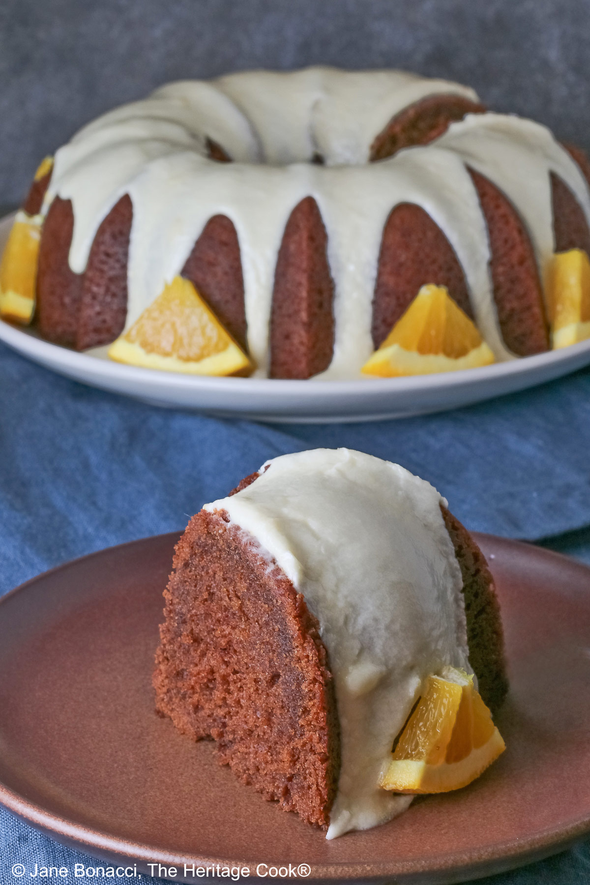 Whole chocolate/orange bundt cake with cascading orange flavored glaze and fresh orange slices around the exterior on a white plate, sometimes on its own, sometimes with a slice cut out and on a plate in front, on a blue cloth with a dark gray background © 2026 Jane Bonacci, The Heritage Cook.