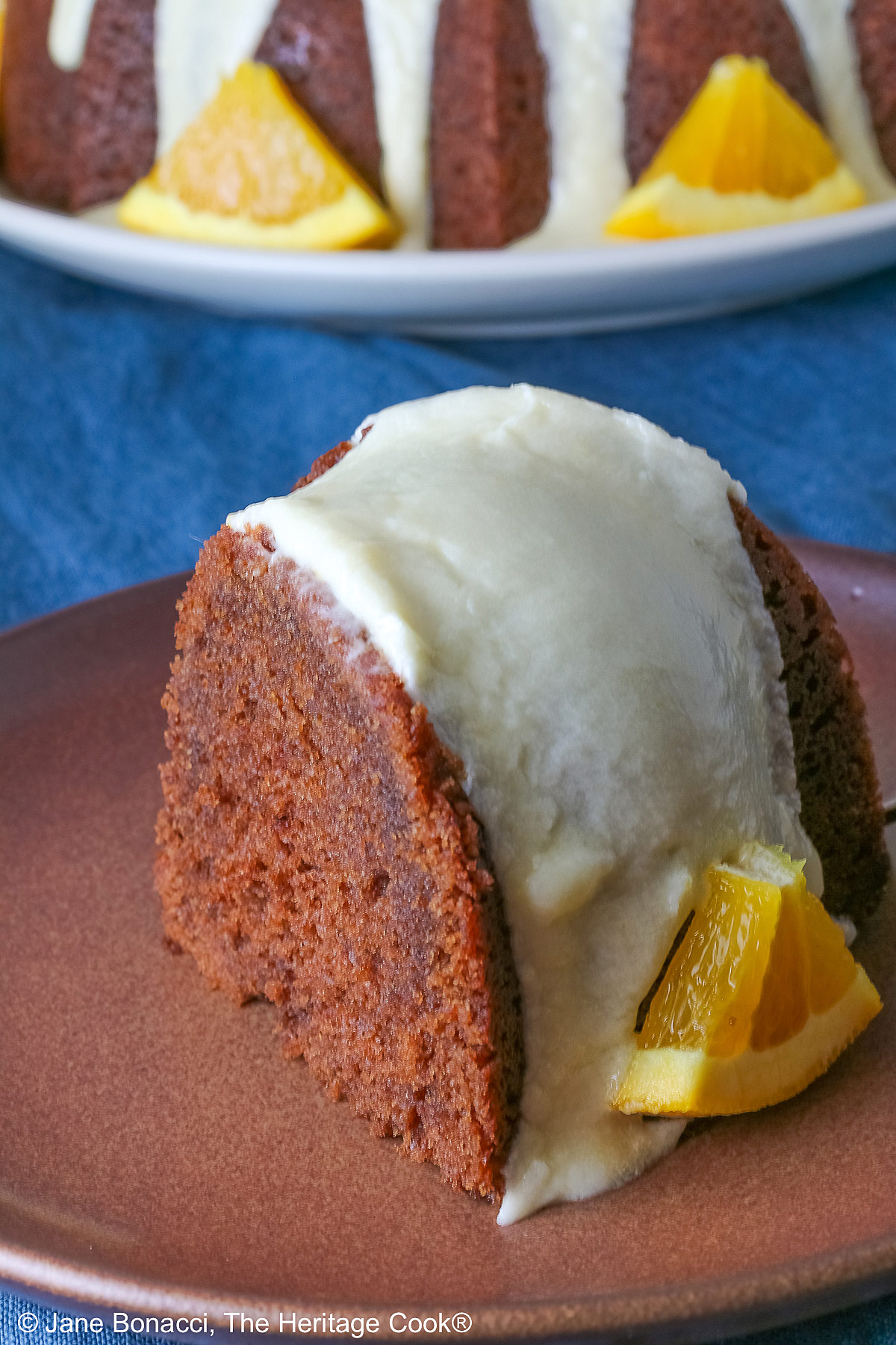 Whole chocolate/orange bundt cake with cascading orange flavored glaze and fresh orange slices around the exterior on a white plate, sometimes on its own, sometimes with a slice cut out and on a plate in front, on a blue cloth with a dark gray background © 2026 Jane Bonacci, The Heritage Cook.