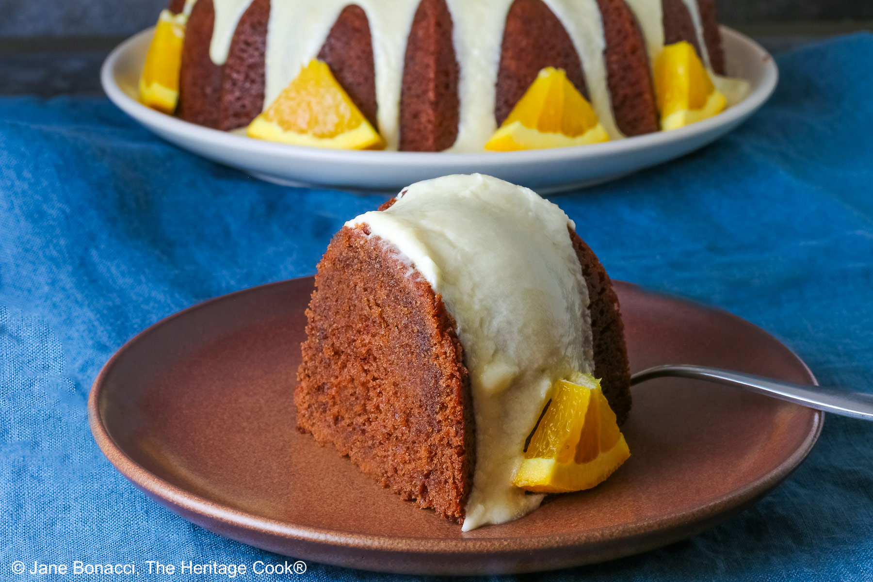 Whole chocolate/orange bundt cake with cascading orange flavored glaze and fresh orange slices around the exterior on a white plate, sometimes on its own, sometimes with a slice cut out and on a plate in front, on a blue cloth with a dark gray background © 2026 Jane Bonacci, The Heritage Cook.