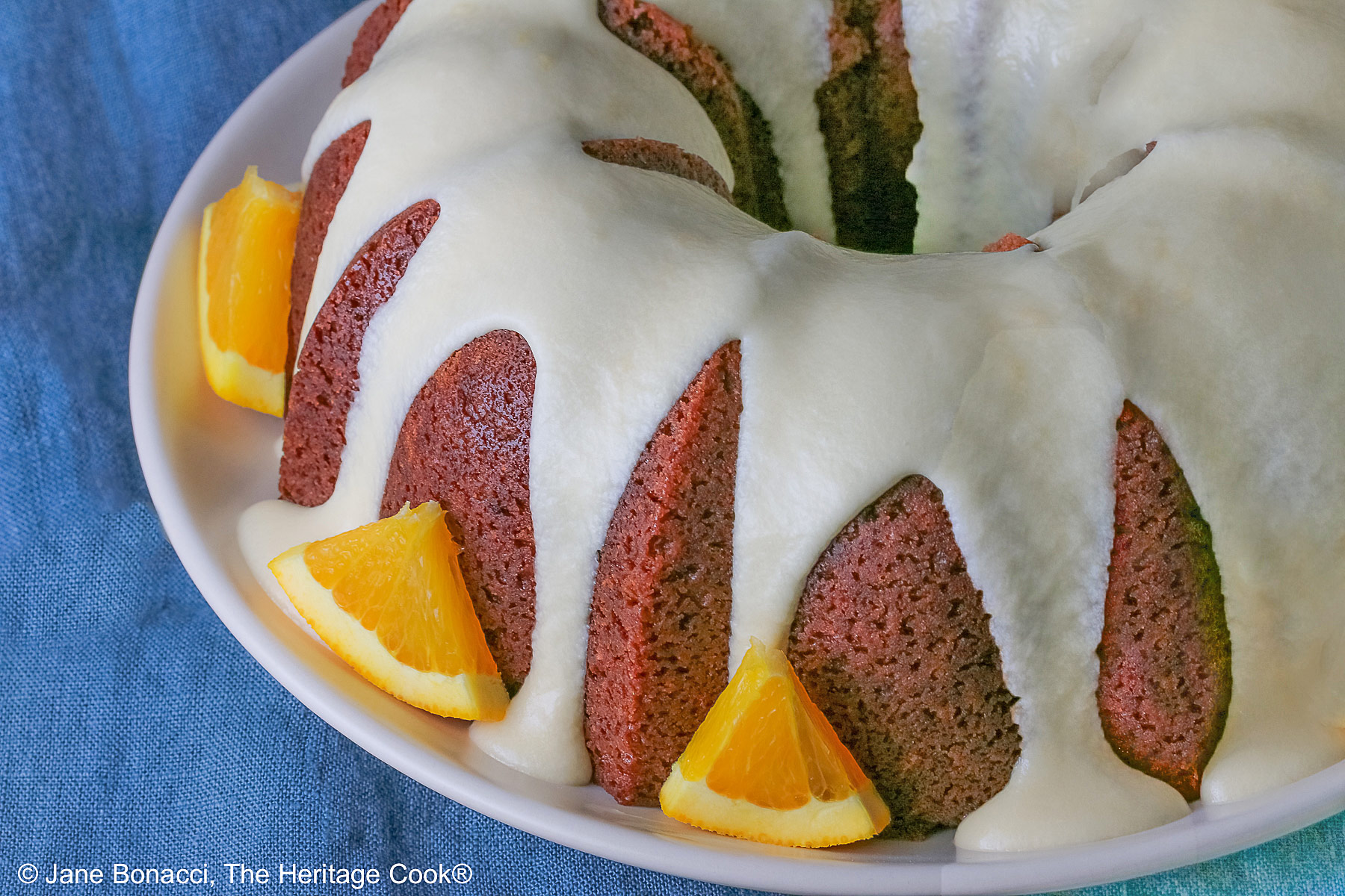 Whole chocolate/orange bundt cake with cascading orange flavored glaze and fresh orange slices around the exterior on a white plate, sometimes on its own, sometimes with a slice cut out and on a plate in front, on a blue cloth with a dark gray background © 2026 Jane Bonacci, The Heritage Cook.