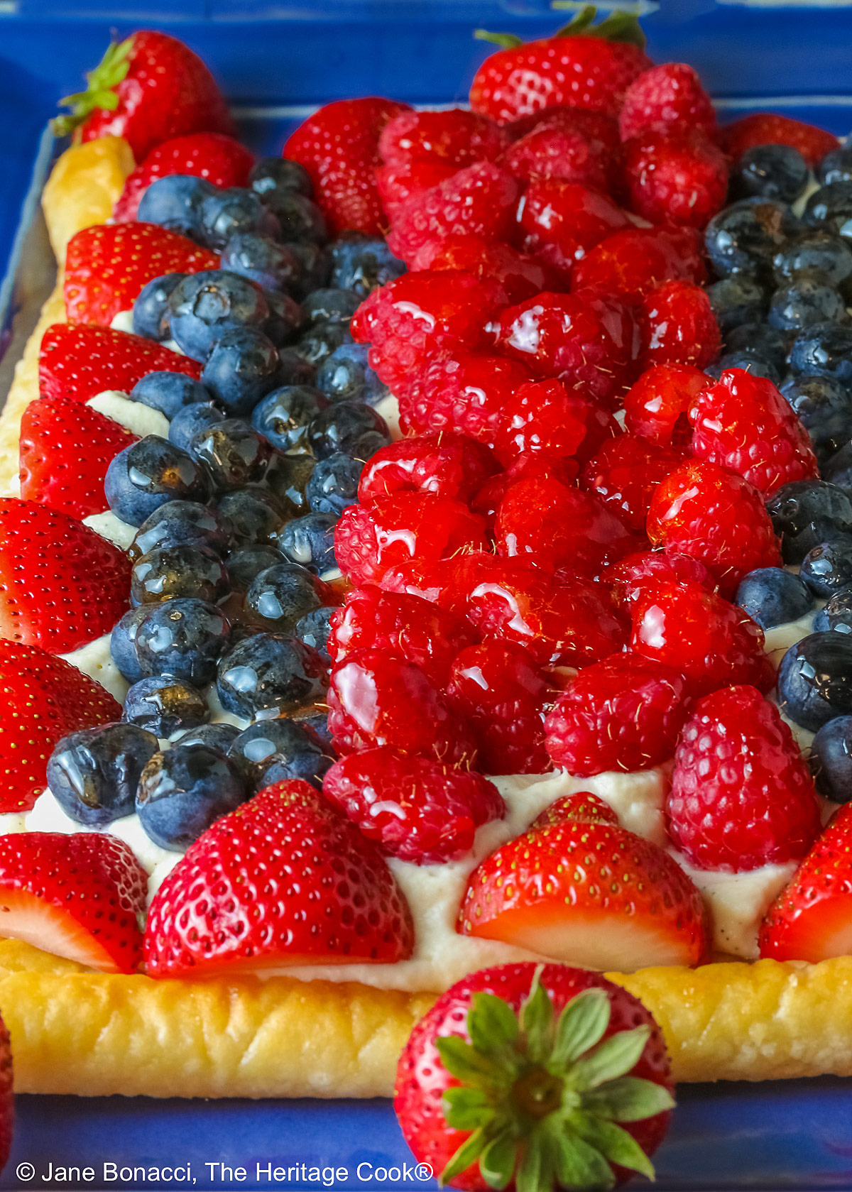 This Patriotic Fruit Tart is sitting on a deep blue platter, with lines of red strawberries, blueberries, and raspberries on a white chocolate cream filling for a fun holiday dessert for your Memorial Day or 4th of July celebrations; some shots of a single slice on a white plate, ready for eating © 2026 Jane Bonacci, The Heritage Cook.