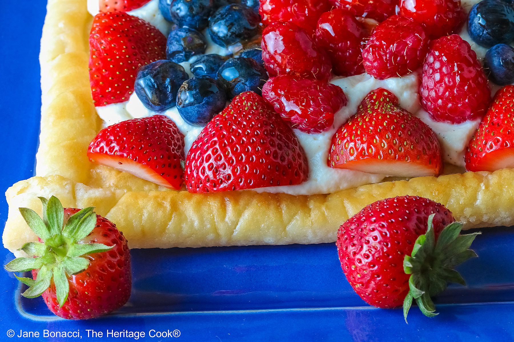 This Patriotic Fruit Tart is sitting on a deep blue platter, with lines of red strawberries, blueberries, and raspberries on a white chocolate cream filling for a fun holiday dessert for your Memorial Day or 4th of July celebrations; some shots of a single slice on a white plate, ready for eating © 2026 Jane Bonacci, The Heritage Cook.