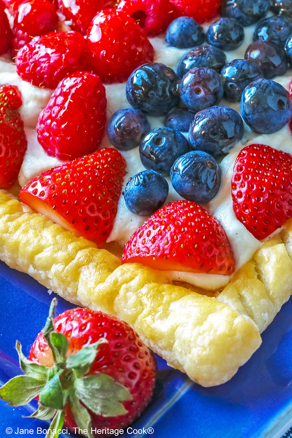 This Patriotic Fruit Tart is sitting on a deep blue platter, with lines of red strawberries, blueberries, and raspberries on a white chocolate cream filling for a fun holiday dessert for your Memorial Day or 4th of July celebrations; some shots of a single slice on a white plate, ready for eating © 2026 Jane Bonacci, The Heritage Cook.