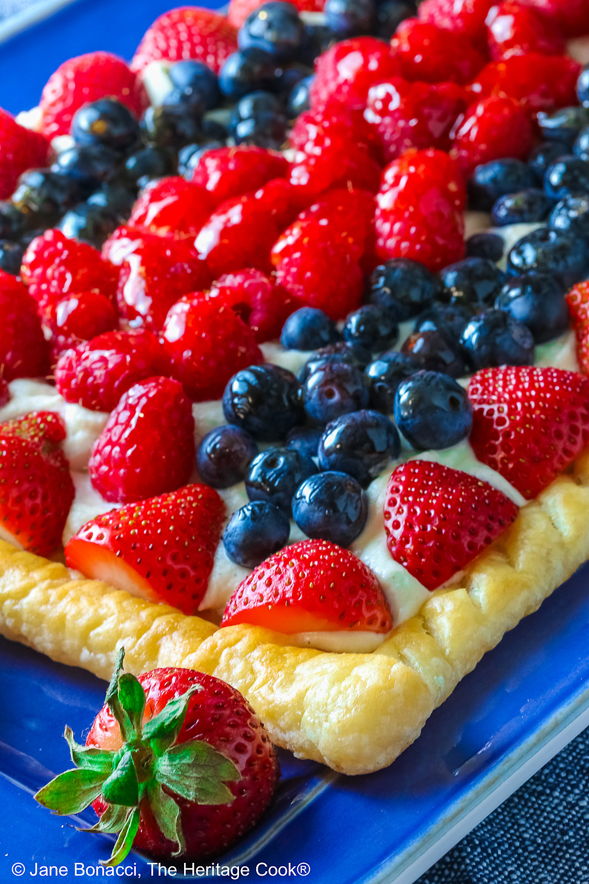 This Patriotic Fruit Tart is sitting on a deep blue platter, with lines of red strawberries, blueberries, and raspberries on a white chocolate cream filling for a fun holiday dessert for your Memorial Day or 4th of July celebrations; some shots of a single slice on a white plate, ready for eating © 2026 Jane Bonacci, The Heritage Cook.