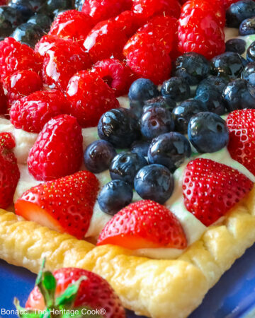 This Patriotic Fruit Tart is sitting on a deep blue platter, with lines of red strawberries, blueberries, and raspberries on a white chocolate cream filling for a fun holiday dessert for your Memorial Day or 4th of July celebrations; some shots of a single slice on a white plate, ready for eating © 2026 Jane Bonacci, The Heritage Cook.