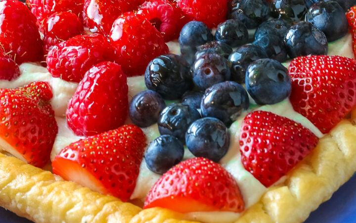 This Patriotic Fruit Tart is sitting on a deep blue platter, with lines of red strawberries, blueberries, and raspberries on a white chocolate cream filling for a fun holiday dessert for your Memorial Day or 4th of July celebrations; some shots of a single slice on a white plate, ready for eating © 2026 Jane Bonacci, The Heritage Cook.
