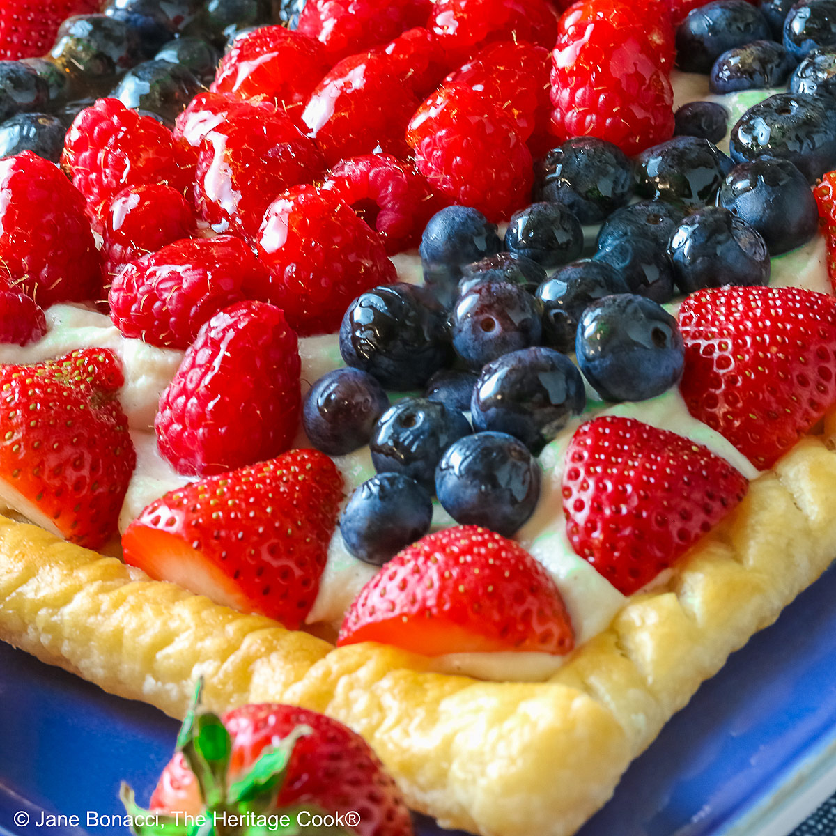 This Patriotic Fruit Tart is sitting on a deep blue platter, with lines of red strawberries, blueberries, and raspberries on a white chocolate cream filling for a fun holiday dessert for your Memorial Day or 4th of July celebrations; some shots of a single slice on a white plate, ready for eating © 2026 Jane Bonacci, The Heritage Cook.