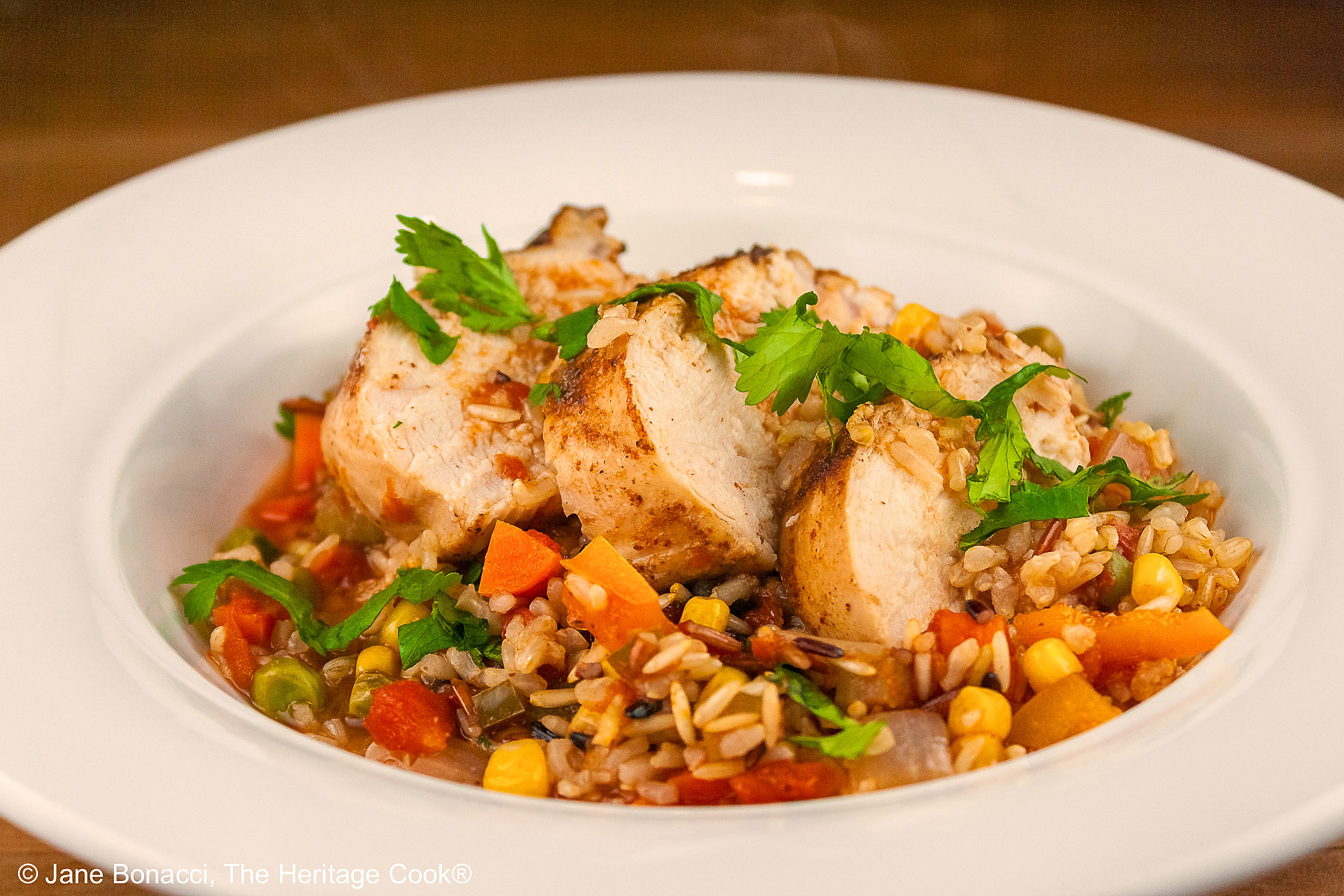 Tex-Mex Arroz con Pollo with colorful rice and vegetables topped with slices of chicken breast and sprinkled with chopped cilantro/parsley in a white bowl with a wide rim © 2026 Jane Bonacci, The Heritage Cook.