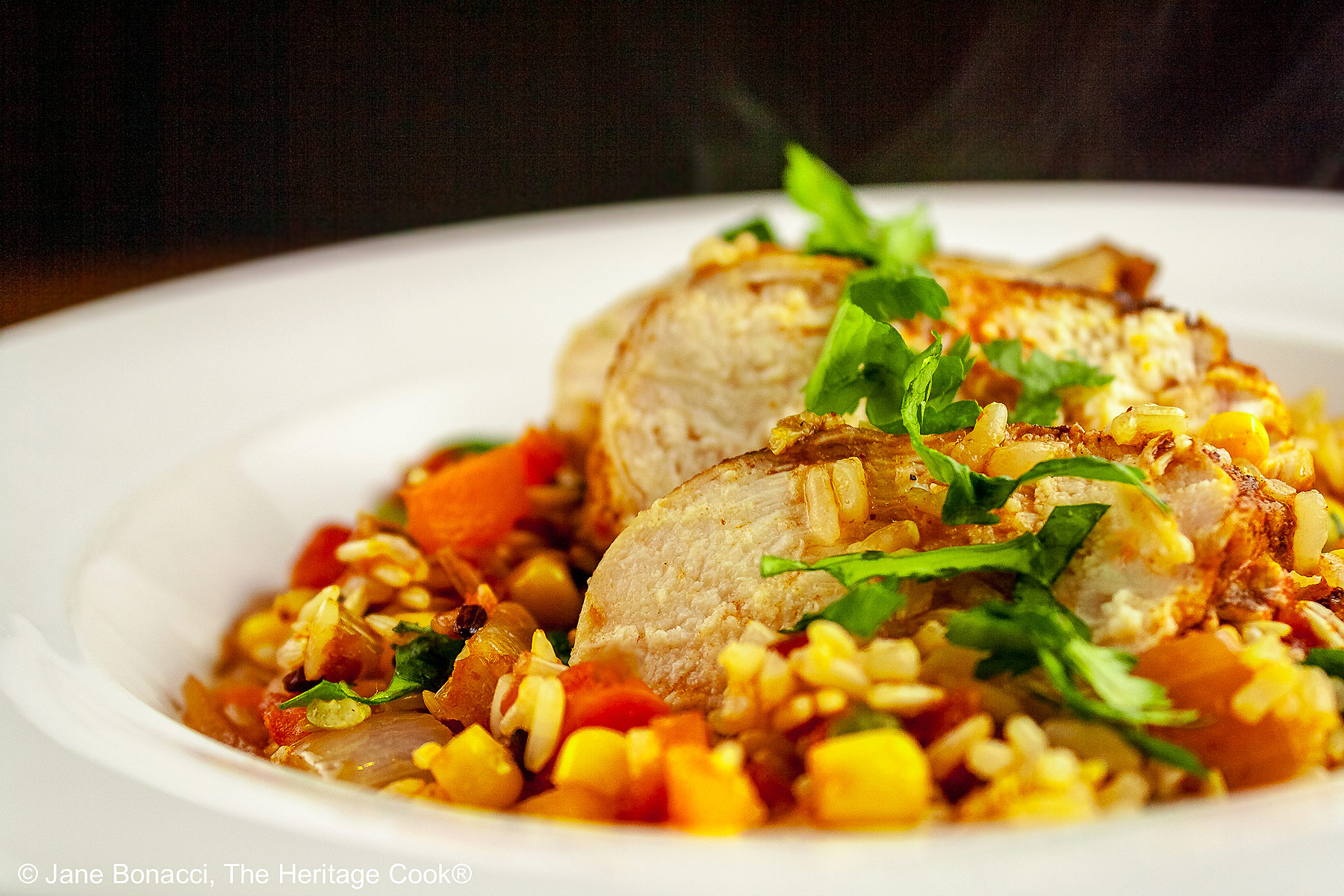 Tex-Mex Arroz con Pollo with colorful rice and vegetables topped with slices of chicken breast and sprinkled with chopped cilantro/parsley in a white bowl with a wide rim © 2026 Jane Bonacci, The Heritage Cook.