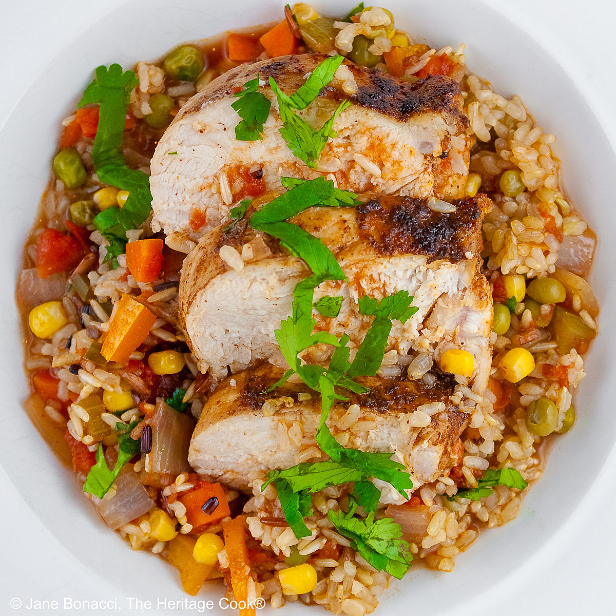 Colorful rice and vegetables with slices of chicken breast laid on top and sprinkled with chopped cilantro/parsley in a white bowl with a wide rim © 2026 Jane Bonacci, The Heritage Cook.