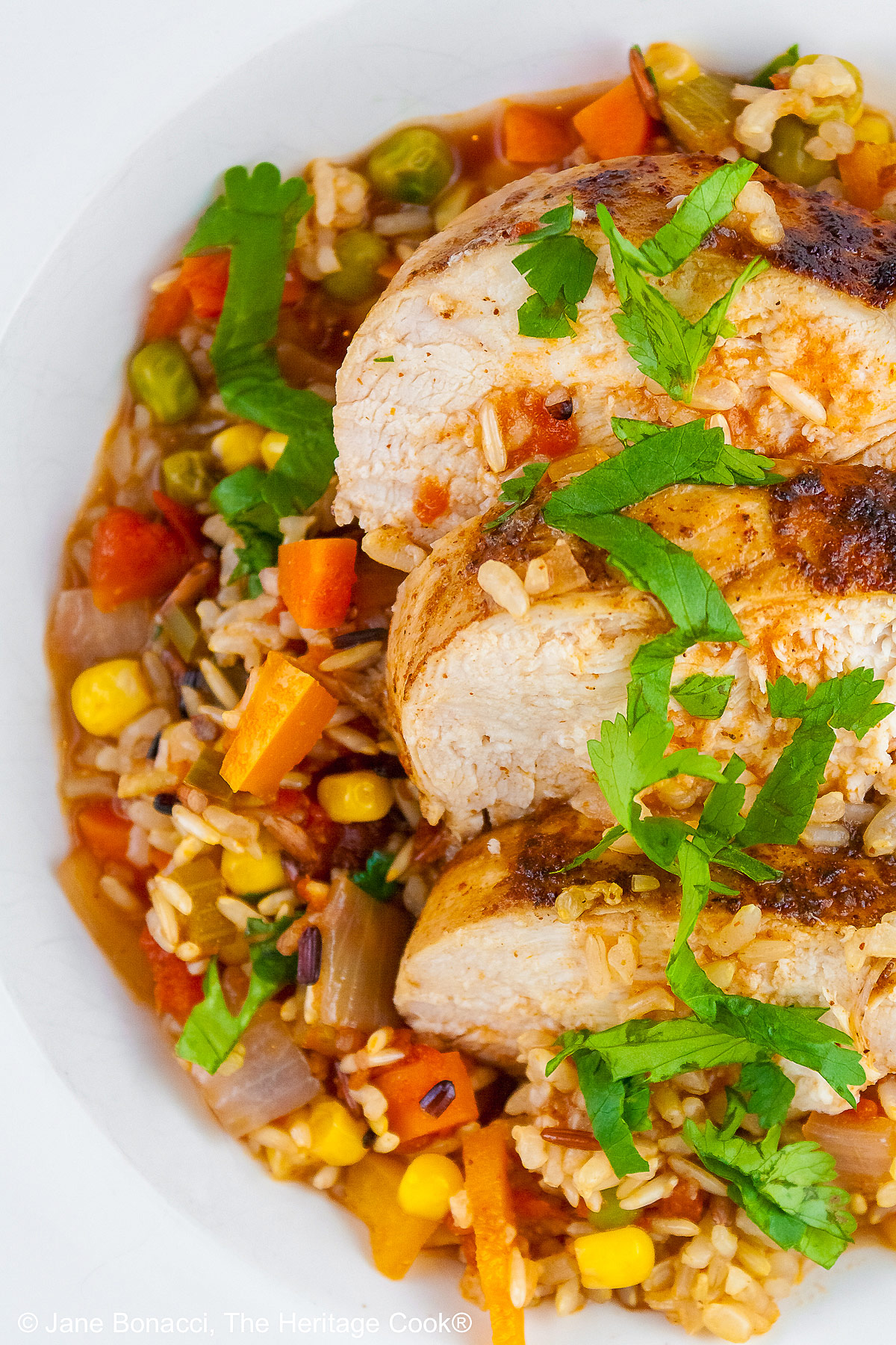 Tex-Mex Arroz con Pollo with colorful rice and vegetables topped with slices of chicken breast and sprinkled with chopped cilantro/parsley in a white bowl with a wide rim © 2026 Jane Bonacci, The Heritage Cook.