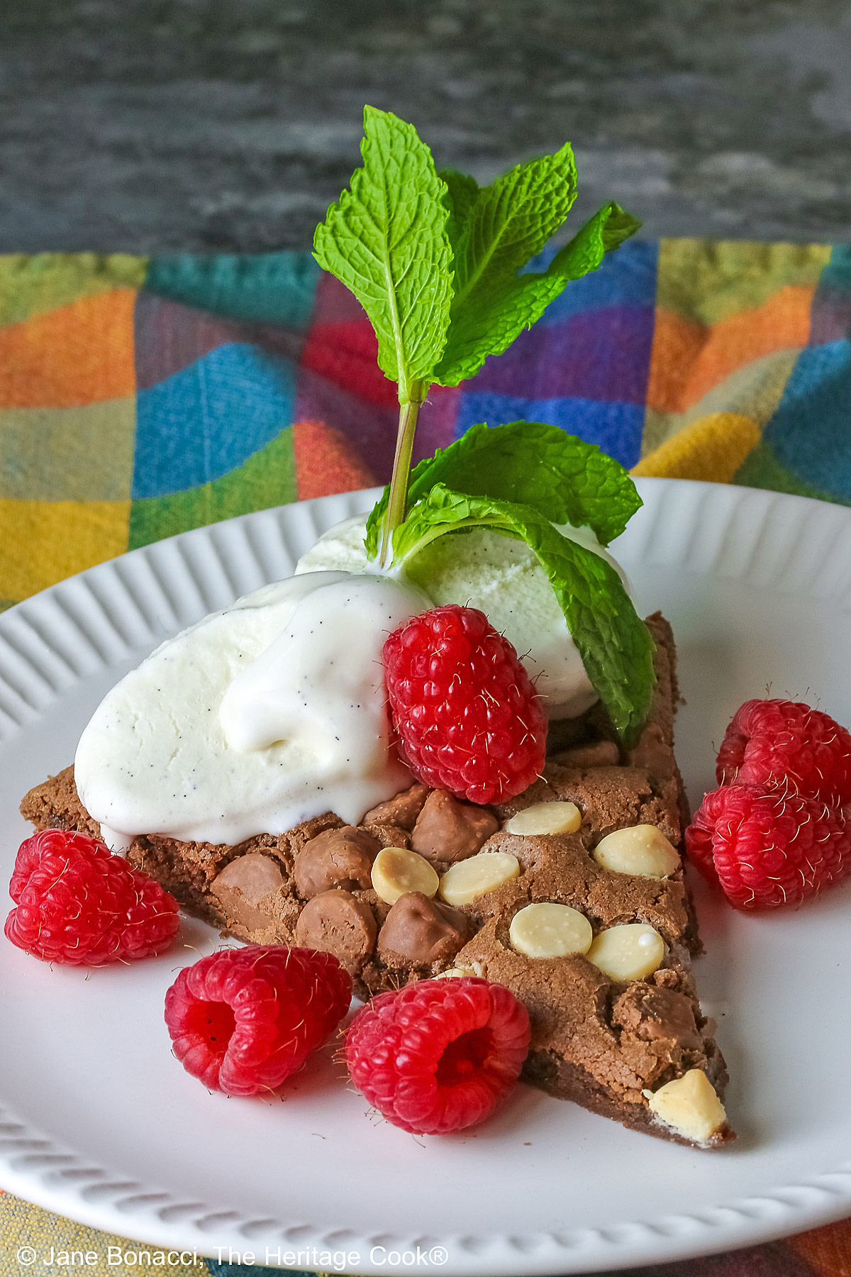 A wedge of the 10-inch (25cm) round Triple Chocolate Jumbo Cookie topped with scoops of vanilla ice cream, fresh red raspberries, and a sprig of mint © 2026 Jane Bonacci, The Heritage Cook.