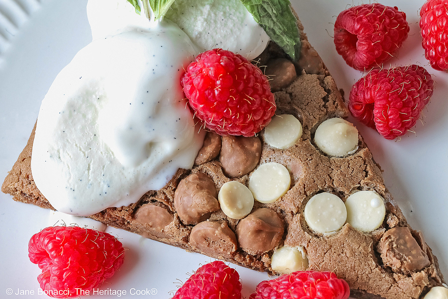 A wedge of the 10-inch (25cm) round Triple Chocolate Jumbo Cookie topped with scoops of vanilla ice cream, fresh red raspberries, and a sprig of mint © 2026 Jane Bonacci, The Heritage Cook.