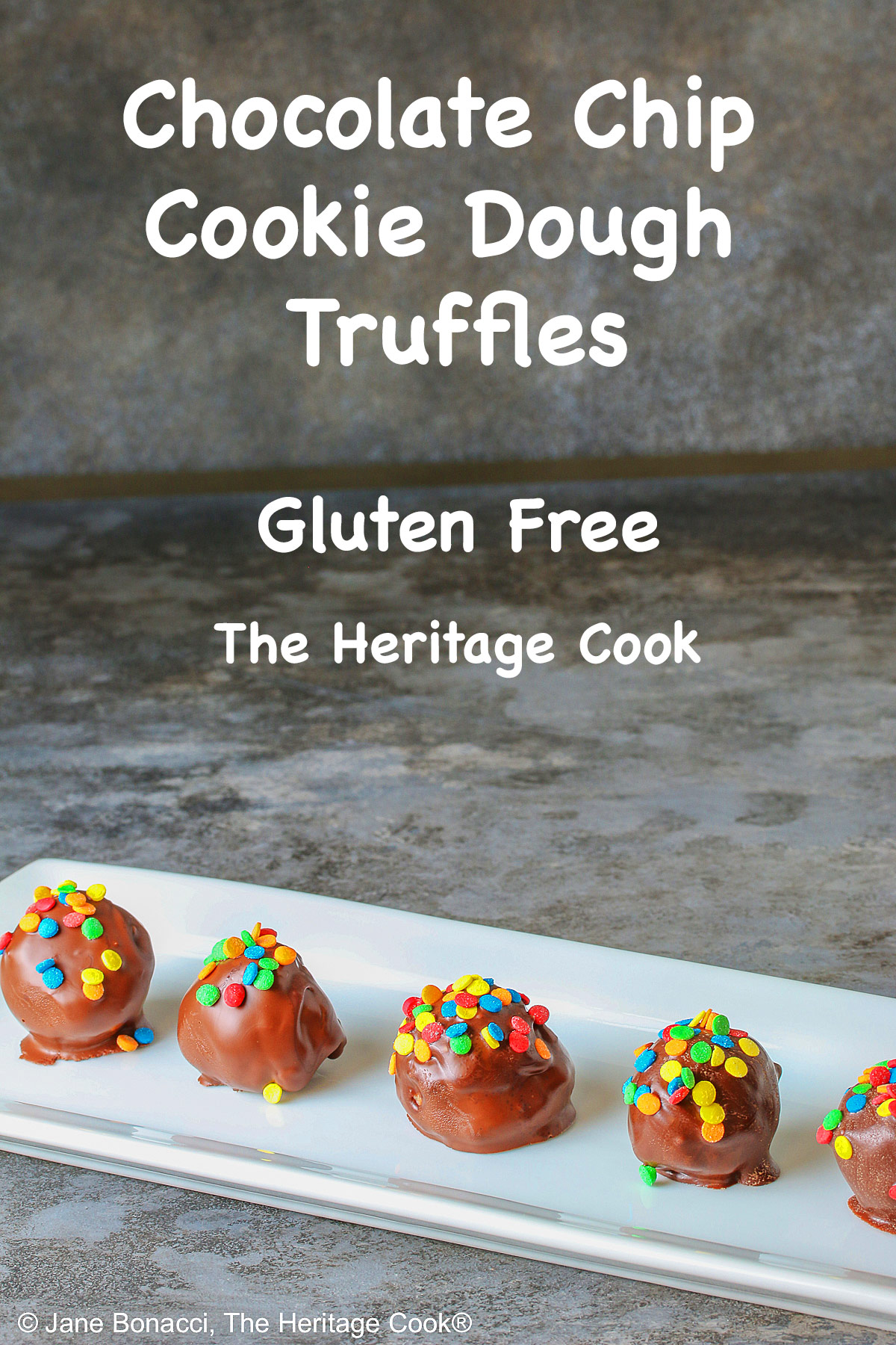 A line of five Chocolate Chip Cookie Dough Truffles on a white rectangular platter, each one dipped in chocolate with brightly colored confetti candy sprinkles on top of each © 2026 Jane Bonacci, The Heritage Cook.