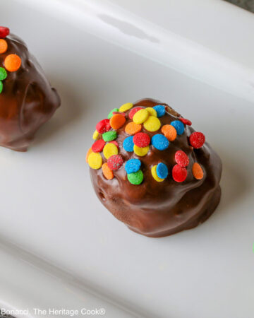 A line of five Chocolate Chip Cookie Dough Truffles on a white rectangular platter, each one dipped in chocolate with brightly colored confetti candy sprinkles on top of each © 2026 Jane Bonacci, The Heritage Cook.
