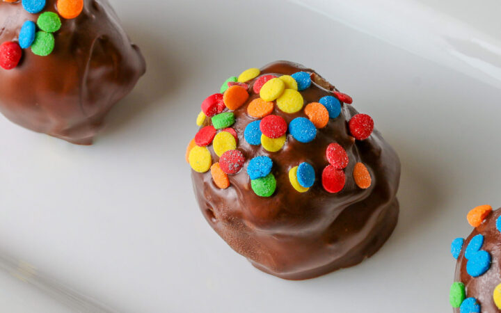 A line of five Chocolate Chip Cookie Dough Truffles on a white rectangular platter, each one dipped in chocolate with brightly colored confetti candy sprinkles on top of each © 2026 Jane Bonacci, The Heritage Cook.