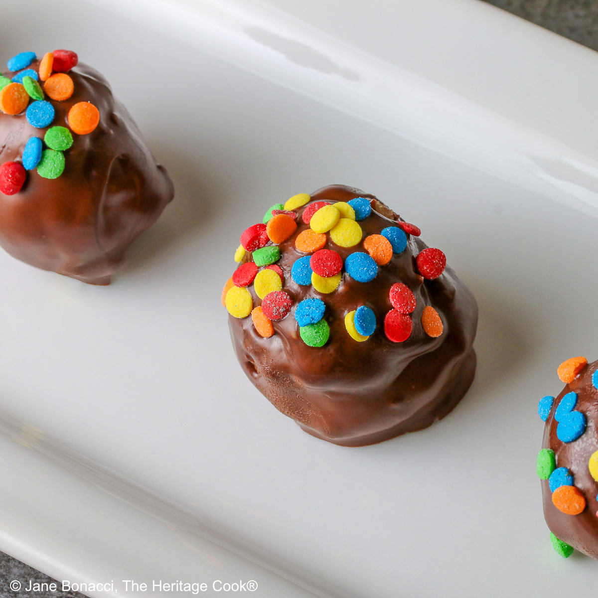 A line of five Chocolate Chip Cookie Dough Truffles on a white rectangular platter, each one dipped in chocolate with brightly colored confetti candy sprinkles on top of each © 2026 Jane Bonacci, The Heritage Cook.