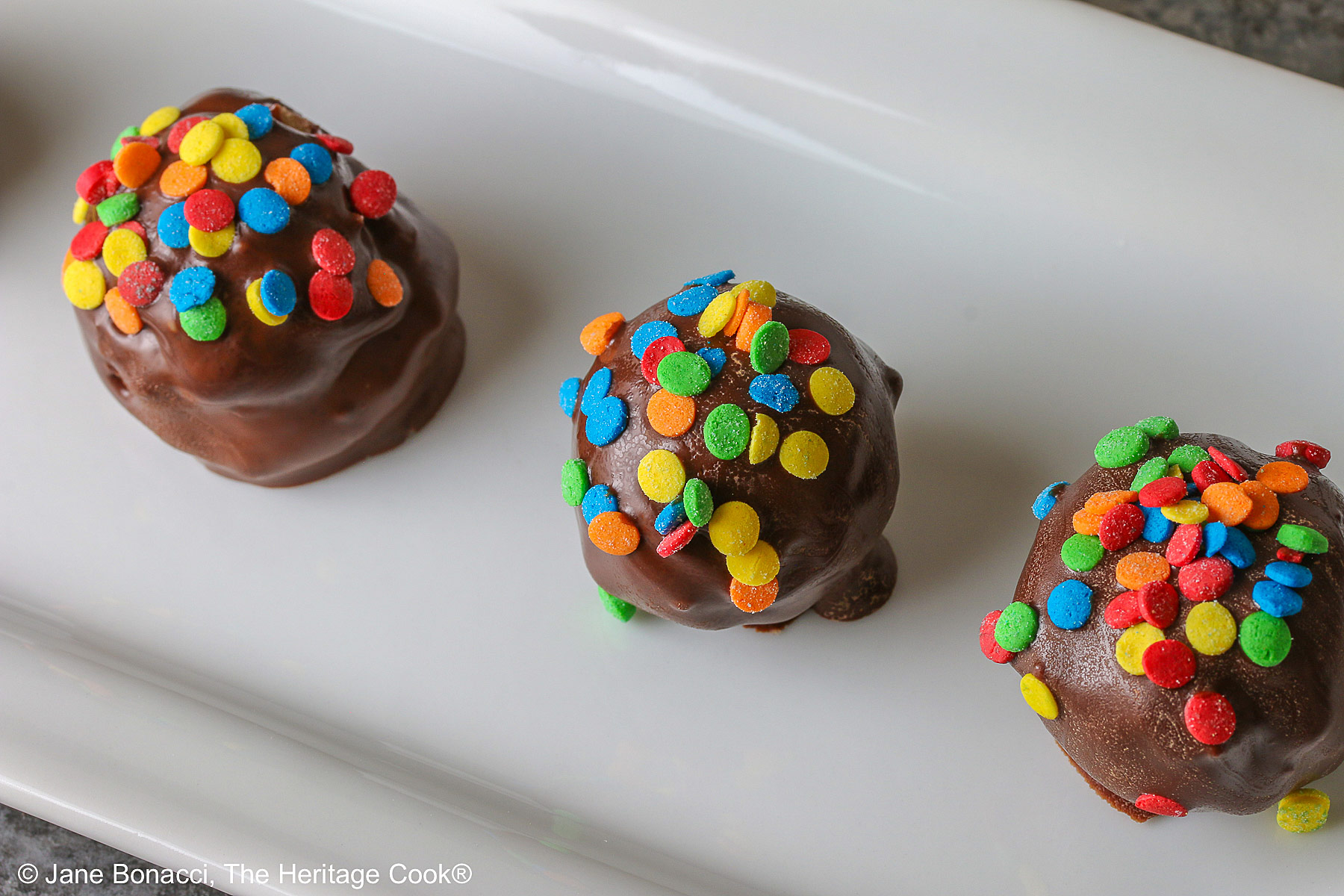 A line of five Chocolate Chip Cookie Dough Truffles on a white rectangular platter, each one dipped in chocolate with brightly colored confetti candy sprinkles on top of each © 2026 Jane Bonacci, The Heritage Cook.